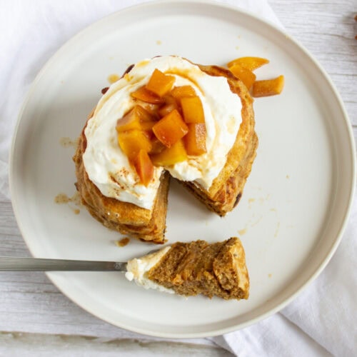 A stack of pancakes topped with whipped cream and fruit compote on a plate, with a slice cut out and resting on a fork. Cinnamon sticks are tied with twine beside the plate.