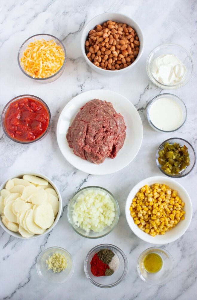 A variety of ingredients in bowls on a marble surface, including ground beef, pinto beans, corn, cheese, potatoes, onions, tomatoes, spices, oil, sour cream, cream, and chopped peppers.