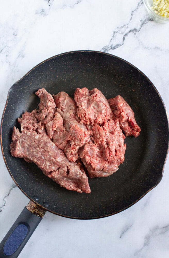 Raw ground beef is placed in a nonstick frying pan on a marble countertop, with a small bowl of minced garlic visible in the corner.