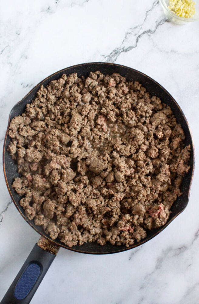 A skillet filled with cooked ground beef sits on a white marble surface, with a small container of minced garlic visible in the corner.