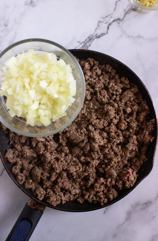 A glass bowl of chopped onions hovers above a pan filled with browned ground beef on a marble countertop.