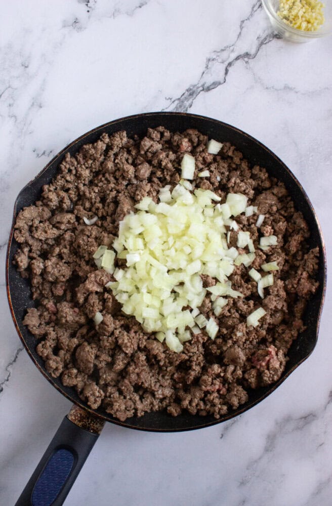 A skillet with cooked ground beef and raw chopped onions in the center, on a marble countertop.