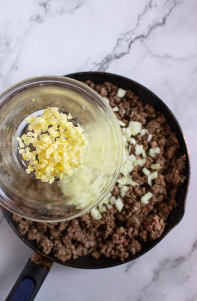 Chopped garlic is being added to a skillet of cooked ground beef and onions on a marble countertop.