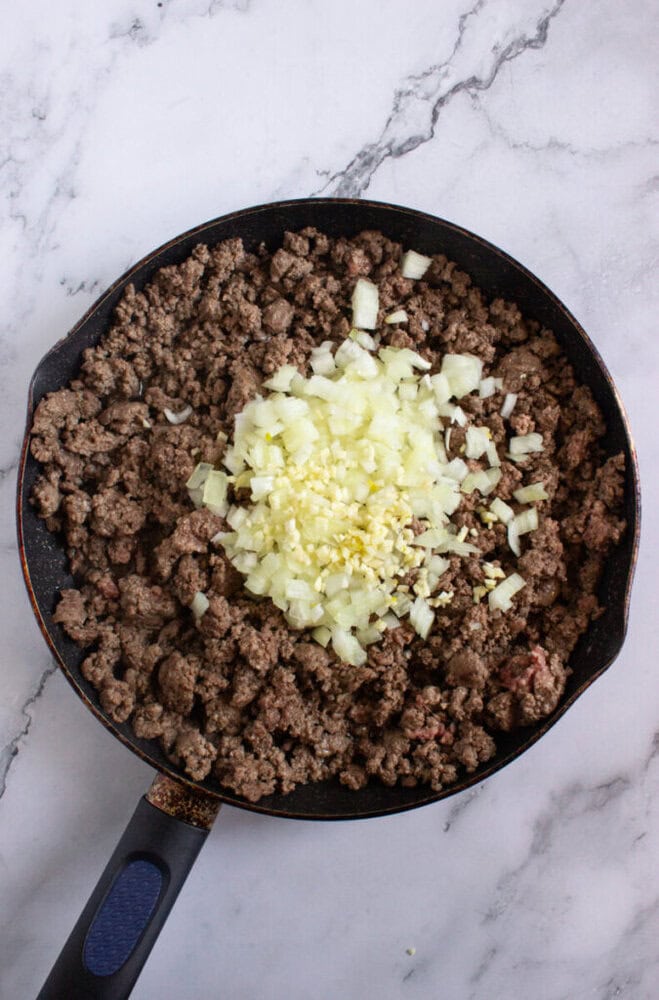 A skillet with cooked ground beef topped with chopped onions and minced garlic, on a marble countertop.