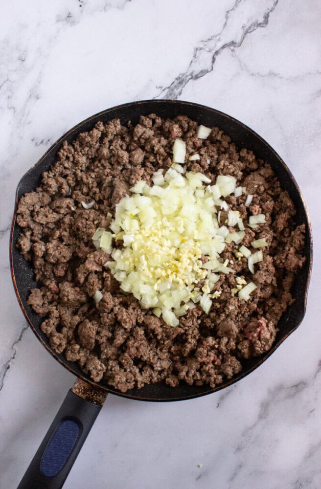 A skillet with cooked ground beef, topped with chopped onions and minced garlic, on a marble countertop.