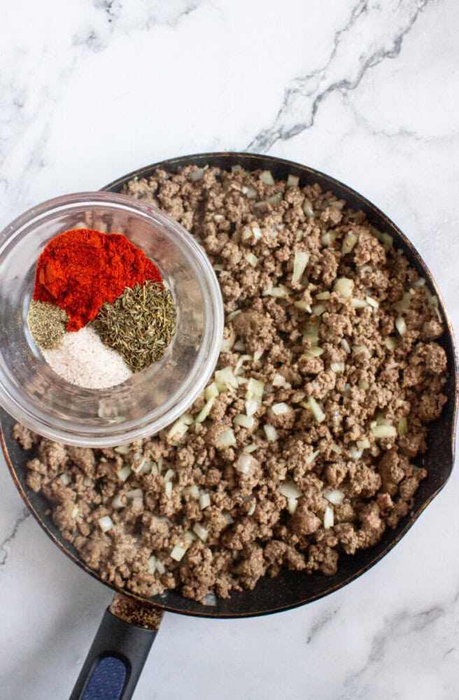 A skillet with cooked ground beef and chopped onions, and a small bowl holding dried herbs and spices placed on top.