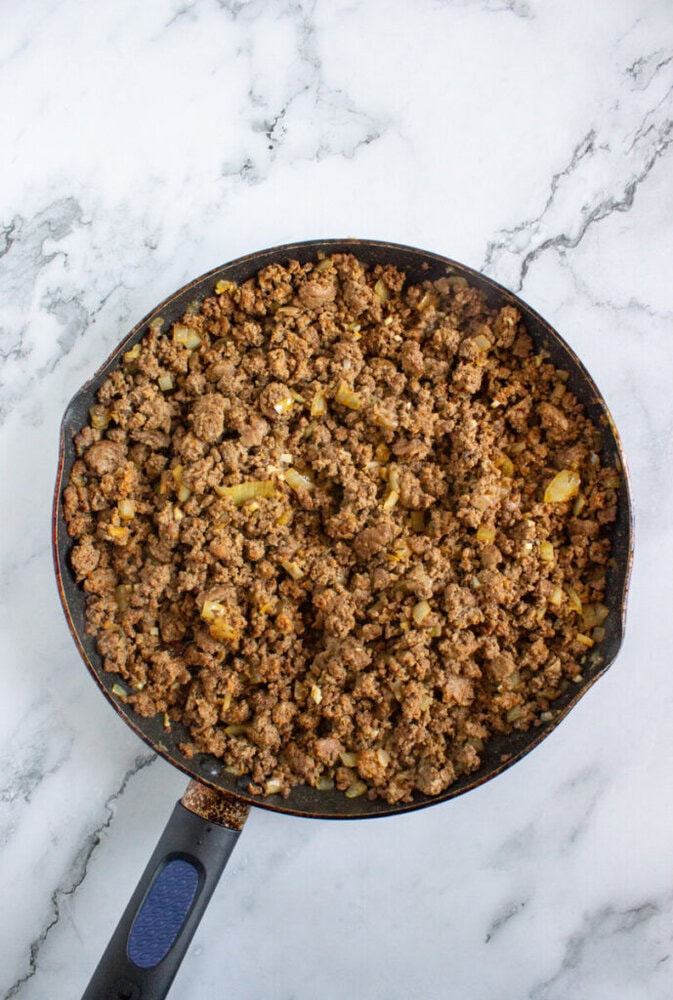 A frying pan filled with cooked ground beef and chopped onions on a marble countertop.