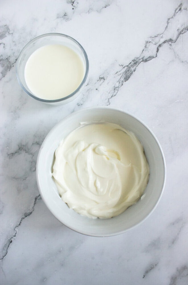 A bowl of thick, white yogurt and a small glass of milk are placed on a light marble surface.