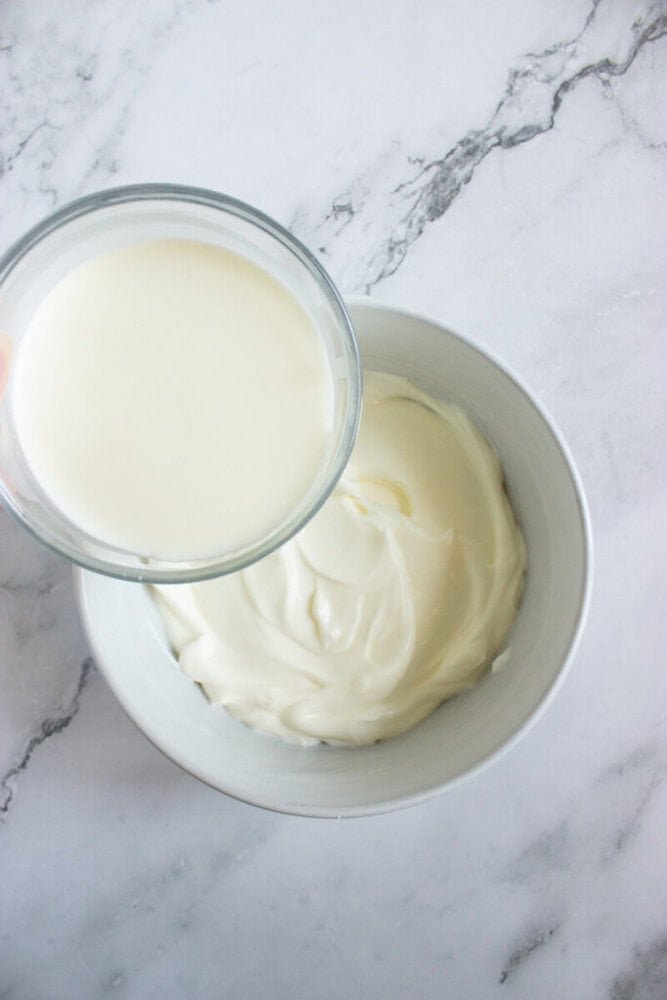 A glass bowl of milk is being held above a white bowl filled with thick, creamy yogurt on a marble surface.