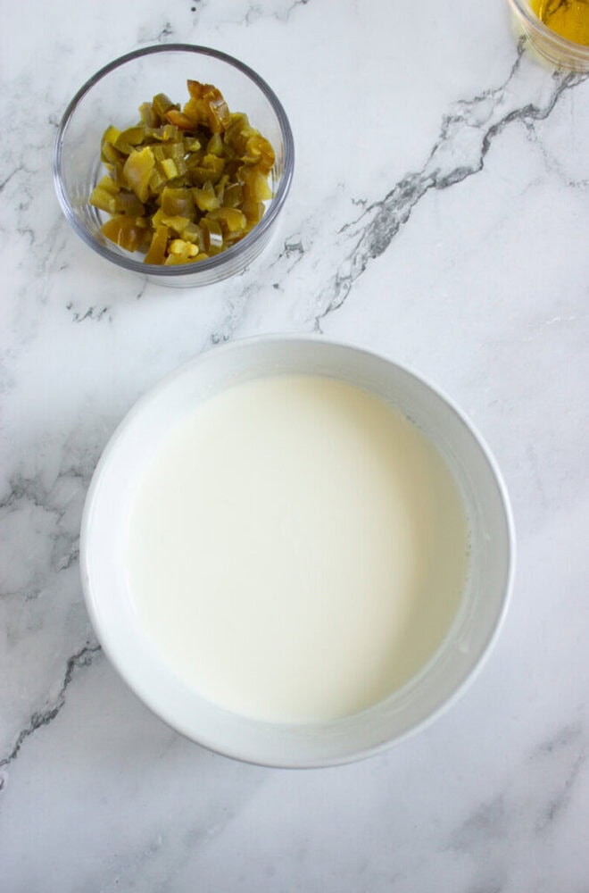 A white bowl filled with a creamy liquid sits on a marble surface next to a small bowl containing chopped pickles.