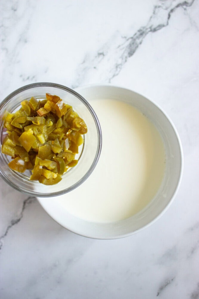 A small glass bowl of chopped pickles next to a white bowl filled with milk, both on a marble surface.