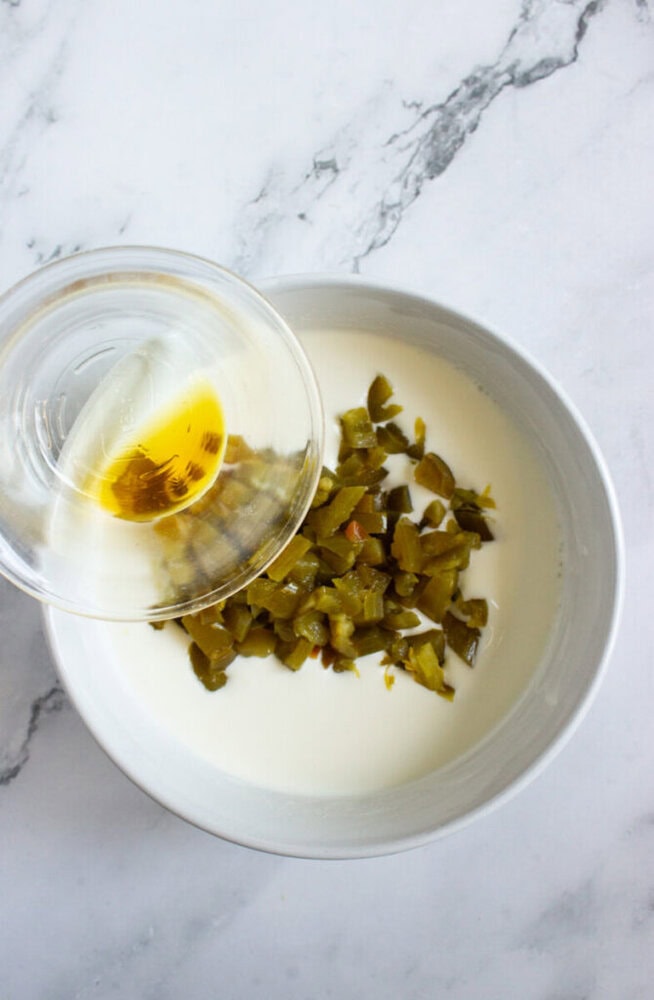 A glass bowl with liquid is being poured into a white bowl containing chopped green chilies and a creamy white substance, on a marble surface.