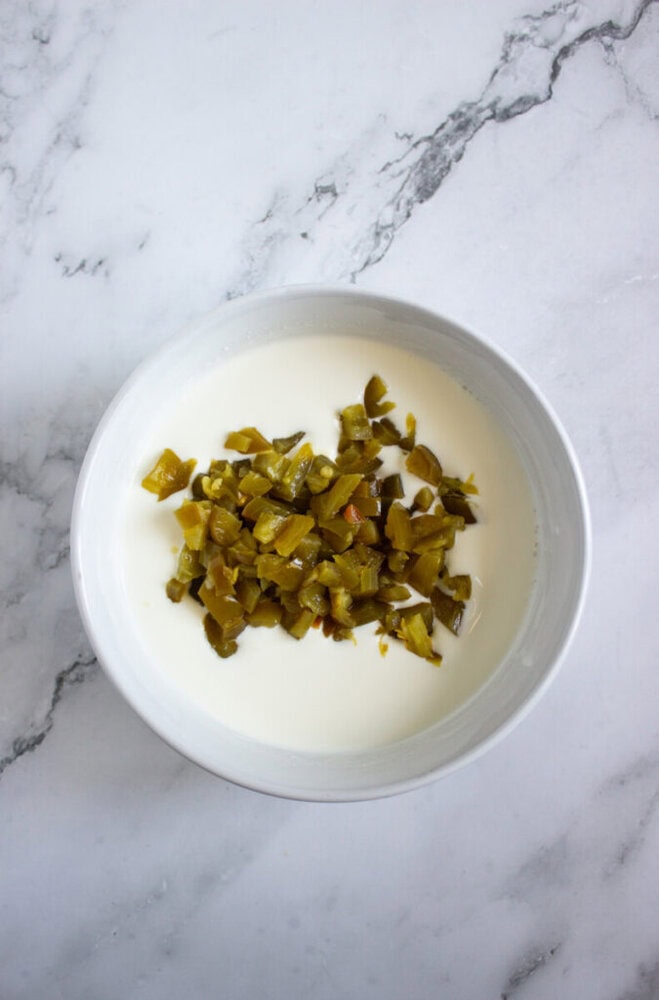 A white bowl containing chopped green chiles in a creamy white liquid, placed on a light marble surface.