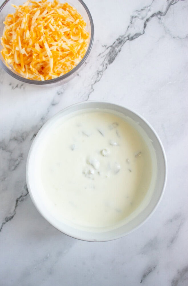 A bowl of creamy white sauce with visible chunks, next to a bowl of shredded yellow and white cheese on a marble surface.