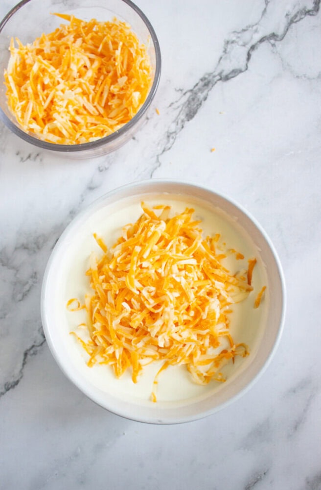 A bowl of shredded cheddar and mozzarella cheese sits on top of milk in a white bowl, with an additional bowl of shredded cheese nearby on a marble surface.
