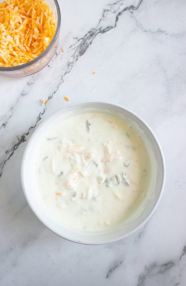 A white bowl containing creamy dip with visible vegetable and cheese pieces sits on a marble surface; shredded cheese is in a separate bowl nearby.