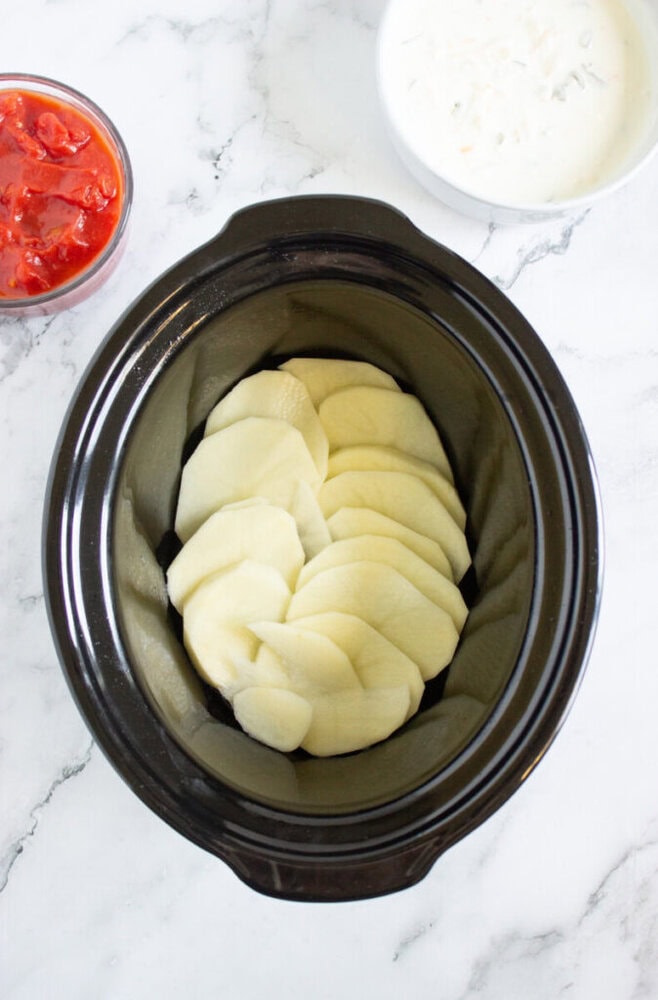A slow cooker with a layer of sliced potatoes inside, next to bowls of diced tomatoes and a creamy white sauce on a marble surface.