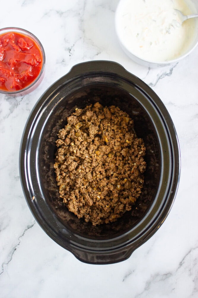 A slow cooker filled with cooked ground beef sits on a marble surface, with bowls of diced tomatoes and shredded cheese nearby.