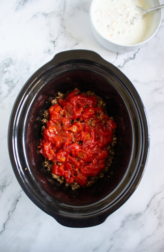 A black slow cooker with cooked ground beef and diced tomatoes, next to a white bowl of creamy white sauce on a marble countertop.