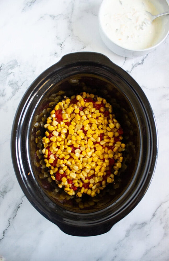 A black slow cooker filled with corn and diced tomatoes sits on a white marble surface, with a bowl of creamy white mixture and a spoon nearby.