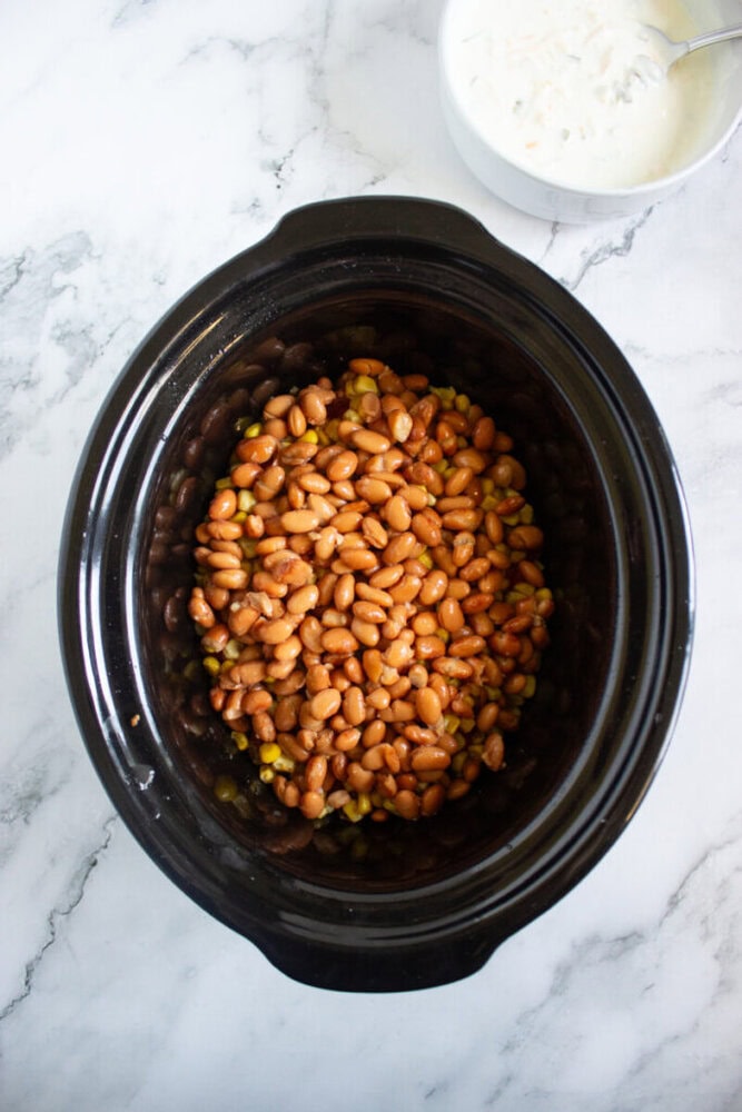 A slow cooker filled with pinto beans and corn sits on a marble countertop, with a bowl of shredded cheese nearby.