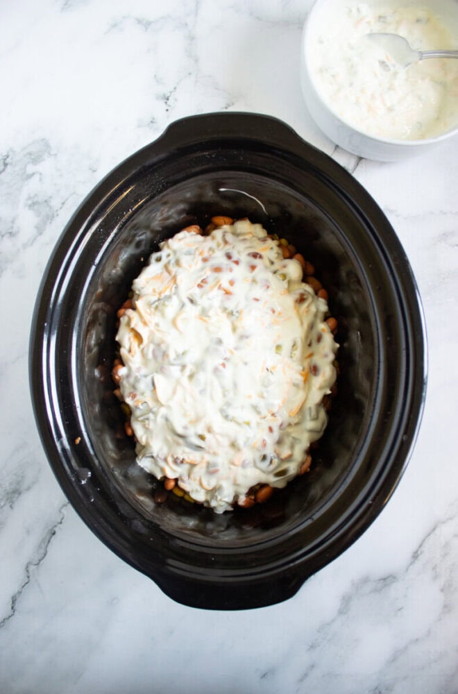 A slow cooker filled with a layered casserole topped with creamy white sauce, with a bowl of extra sauce and a spoon beside it on a marble surface.