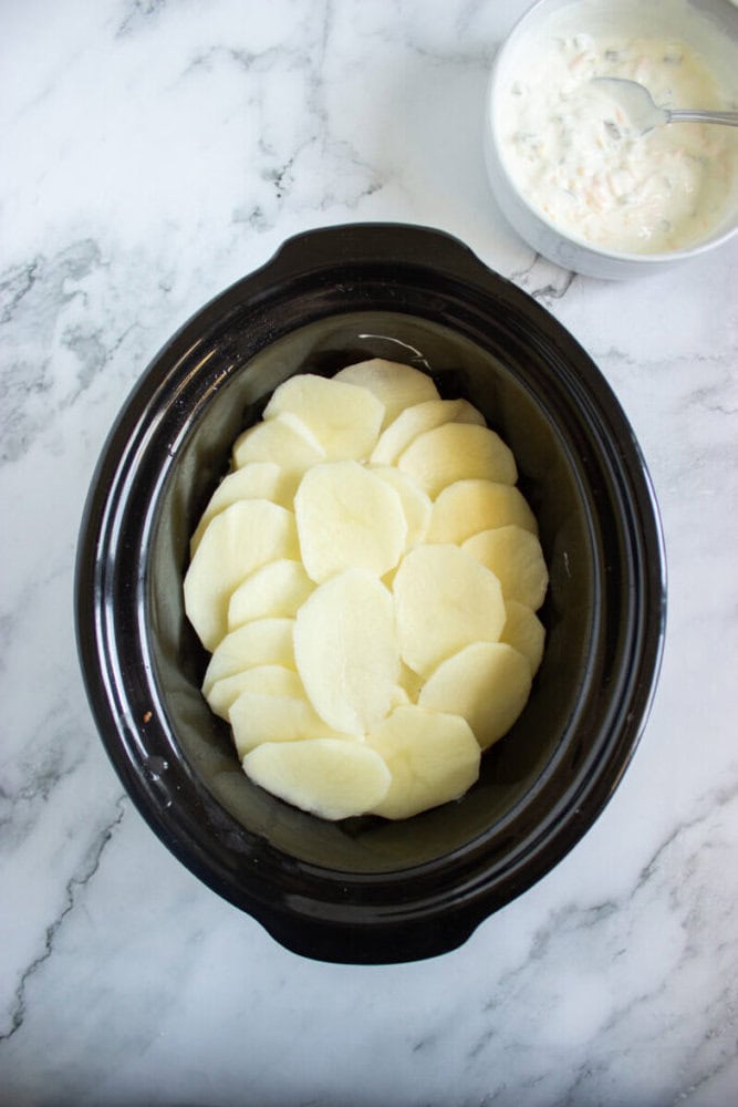Sliced raw potatoes are layered in a black slow cooker, with a bowl of creamy mixture and a spoon beside it on a marble countertop.