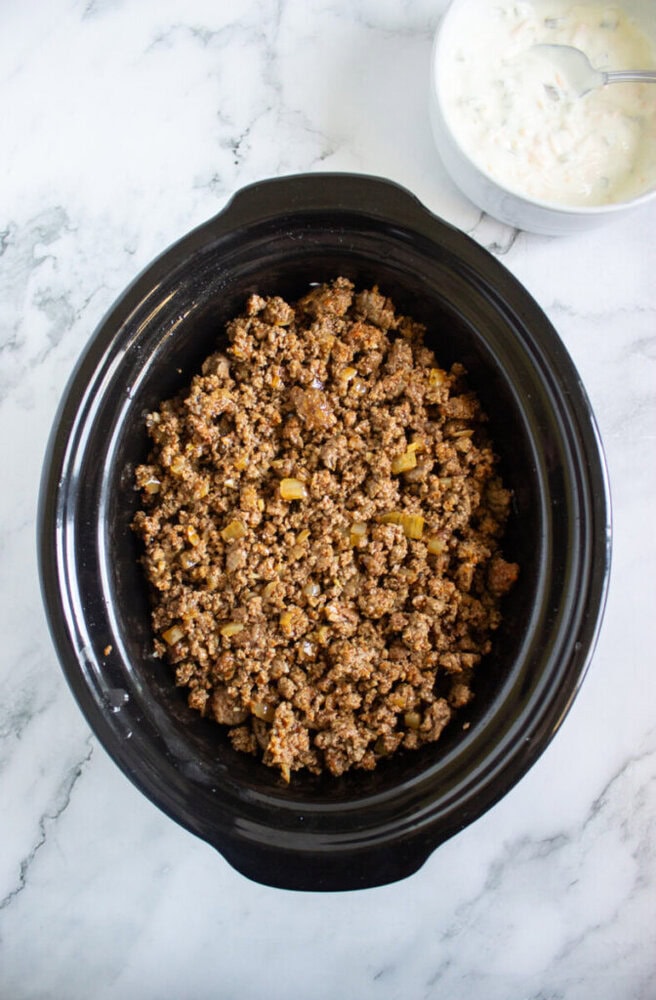 A black slow cooker filled with cooked ground beef mixture sits on a marble surface, with a bowl of white sauce and a spoon nearby.