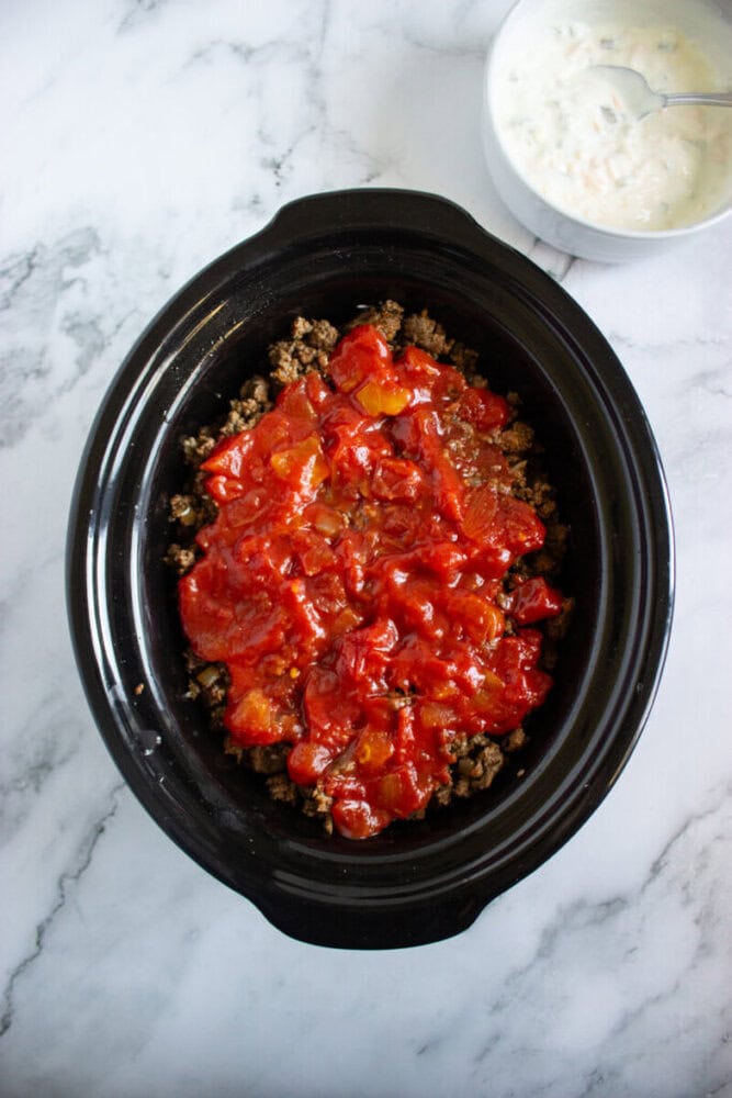 A black slow cooker filled with ground meat and tomatoes sits on a marble surface, with a bowl of white sauce and a spoon nearby.