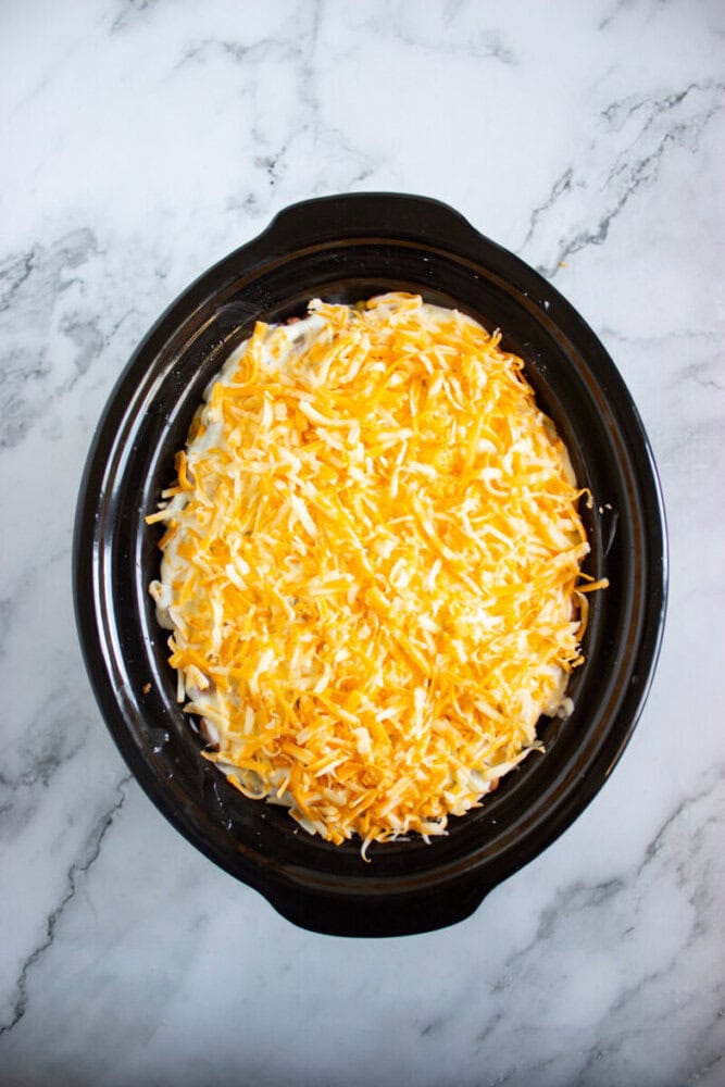 A black slow cooker filled with uncooked macaroni and cheese topped with shredded cheddar cheese, placed on a white marble surface.