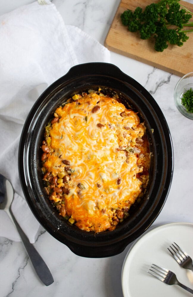A slow cooker filled with a cheesy baked casserole sits on a marble counter, with serving utensils, plates, forks, parsley, and a white napkin nearby.