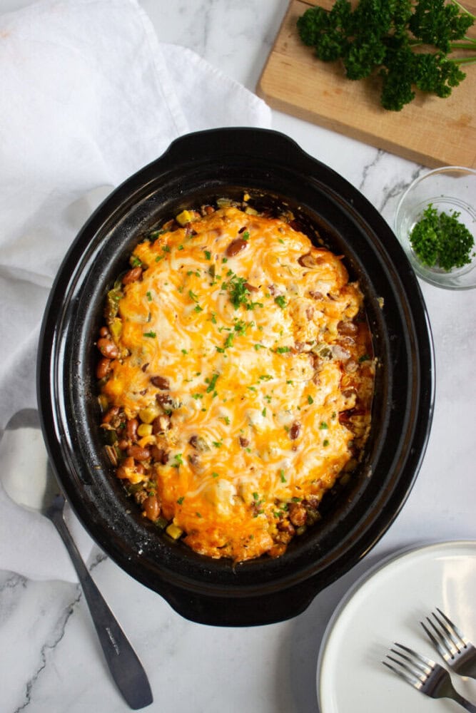 A slow cooker filled with a baked casserole topped with melted cheese and chopped parsley, next to a serving spoon, plates, and a cutting board with fresh herbs.