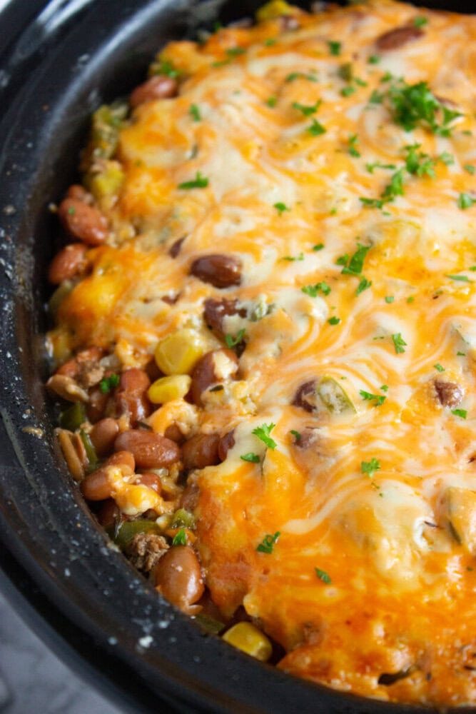 A close-up of a casserole in a slow cooker, featuring melted cheese on top, with visible beans, corn, and ground meat, garnished with chopped parsley.