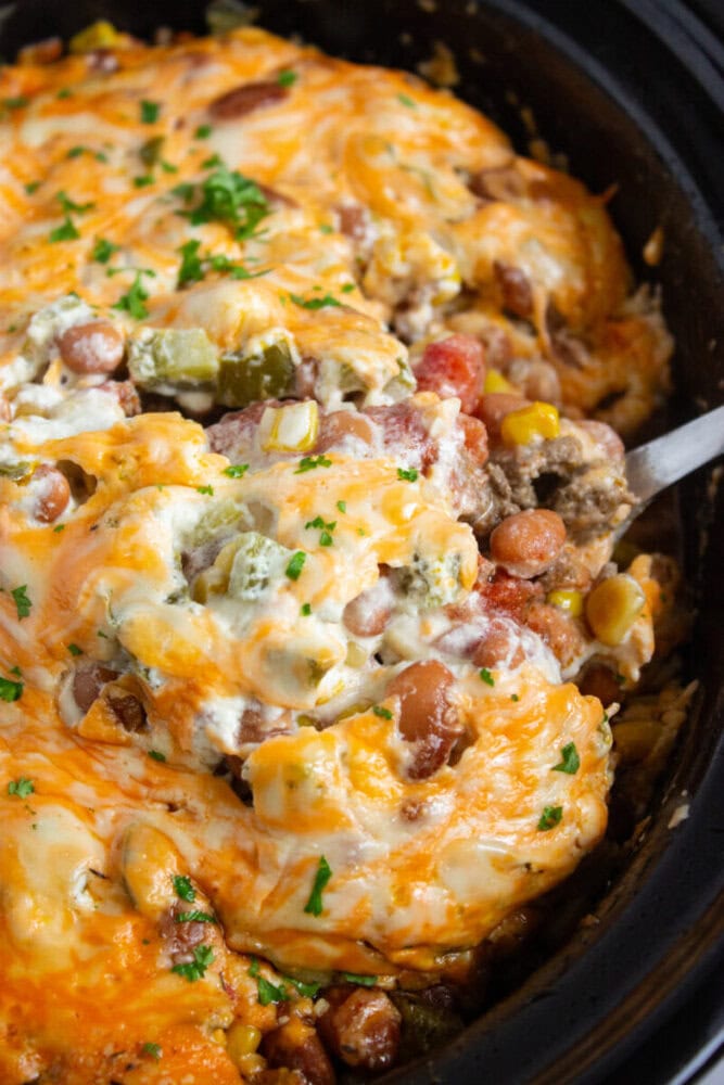 A close-up of a cheesy casserole with beans, corn, ground beef, and green peppers topped with melted cheese and chopped parsley in a black dish.