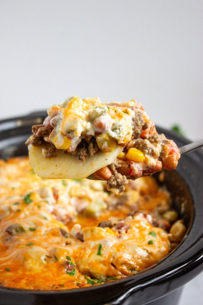 A spoon lifts a serving of cheesy layered casserole with ground beef, beans, corn, and melted cheese from a crockpot.