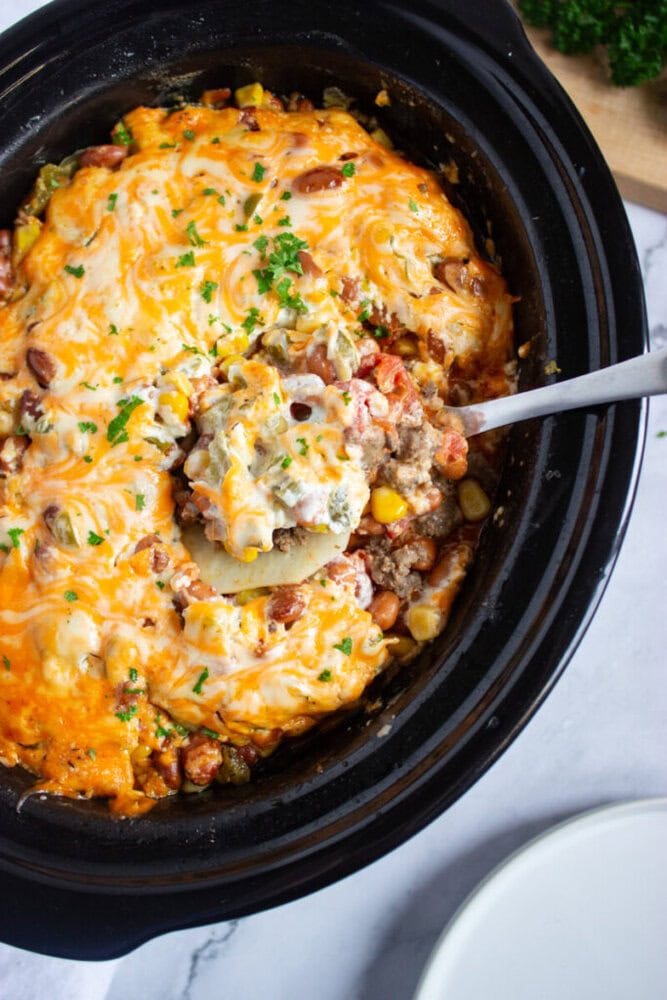 A slow cooker filled with a cheesy casserole containing ground meat, beans, corn, and vegetables; a serving spoon is lifting a portion out.