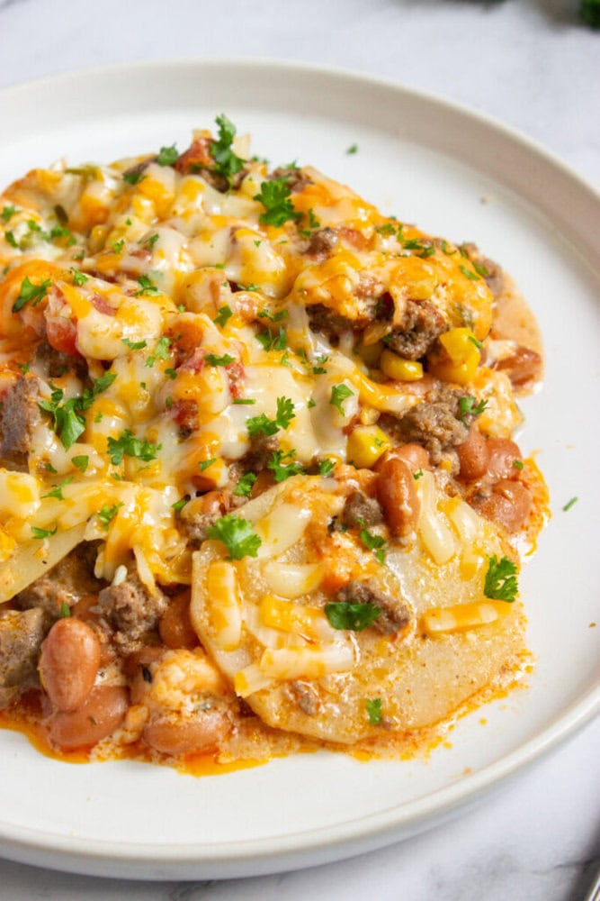 A plate of cheesy casserole topped with melted cheese, ground meat, beans, corn, and chopped parsley.