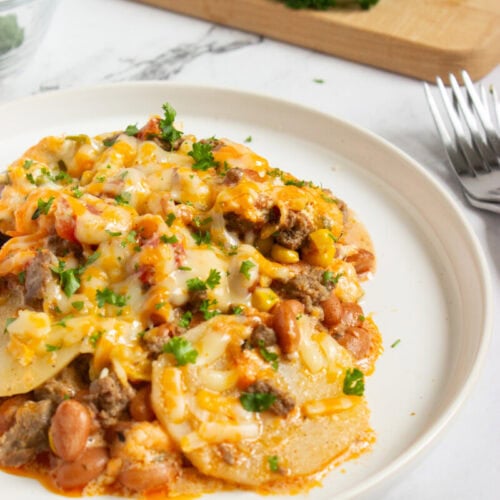 A plate of cheesy casserole with ground meat, beans, corn, and topped with chopped parsley; a cutting board with fresh parsley is in the background.