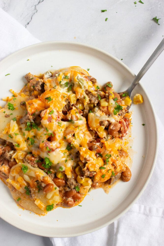 A plate of cheesy casserole with ground meat, corn, beans, tomatoes, and melted cheese, garnished with chopped herbs, served on a white dish with a fork.