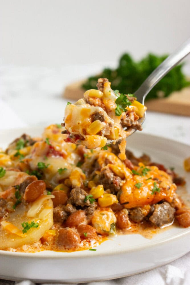 A close-up of a fork holding a cheesy casserole with ground beef, corn, beans, potatoes, and melted cheese, garnished with parsley on a white plate.