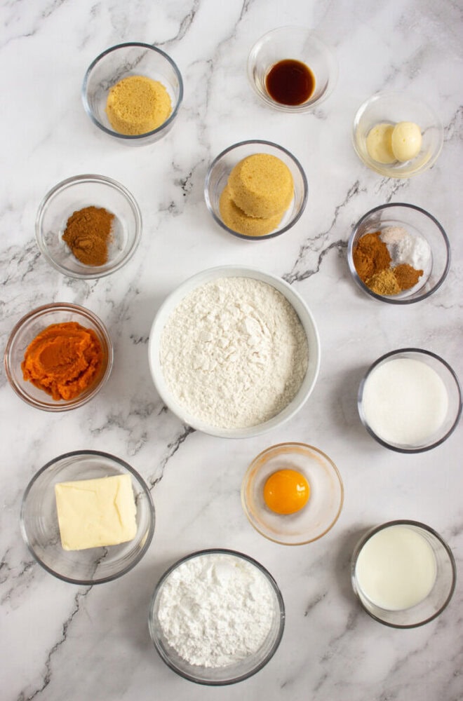 Various baking ingredients, including flour, sugars, pumpkin puree, spices, butter, an egg, milk, vanilla, and baking powder, are arranged in small glass bowls on a marble surface.