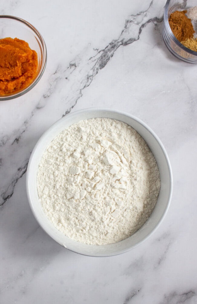 A white bowl filled with flour sits on a marble countertop, with small glass bowls of pumpkin puree and brown sugar nearby.
