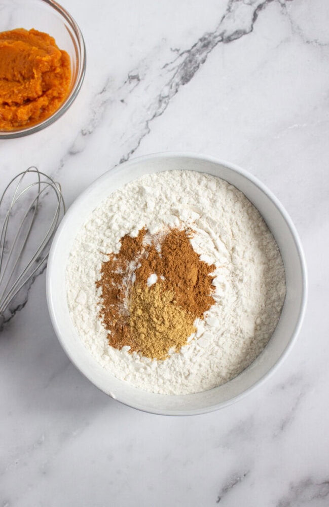 A bowl of flour with ground spices on top sits on a marble surface next to a whisk and a small bowl of orange pumpkin puree.