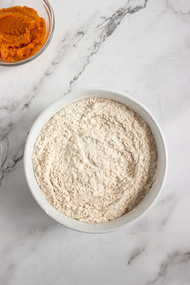 A white bowl filled with flour sits on a marble countertop, with a glass bowl of pumpkin puree partially visible in the upper left corner.