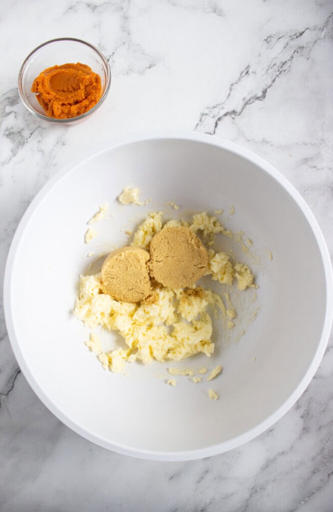 A white mixing bowl contains creamed butter and two scoops of brown sugar. A small glass bowl with pumpkin puree is placed beside the mixing bowl on a marble surface.