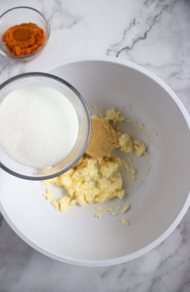 A white bowl with creamed butter and brown sugar, a glass measuring cup with granulated sugar above it, and a small bowl of pumpkin puree on a marble surface.