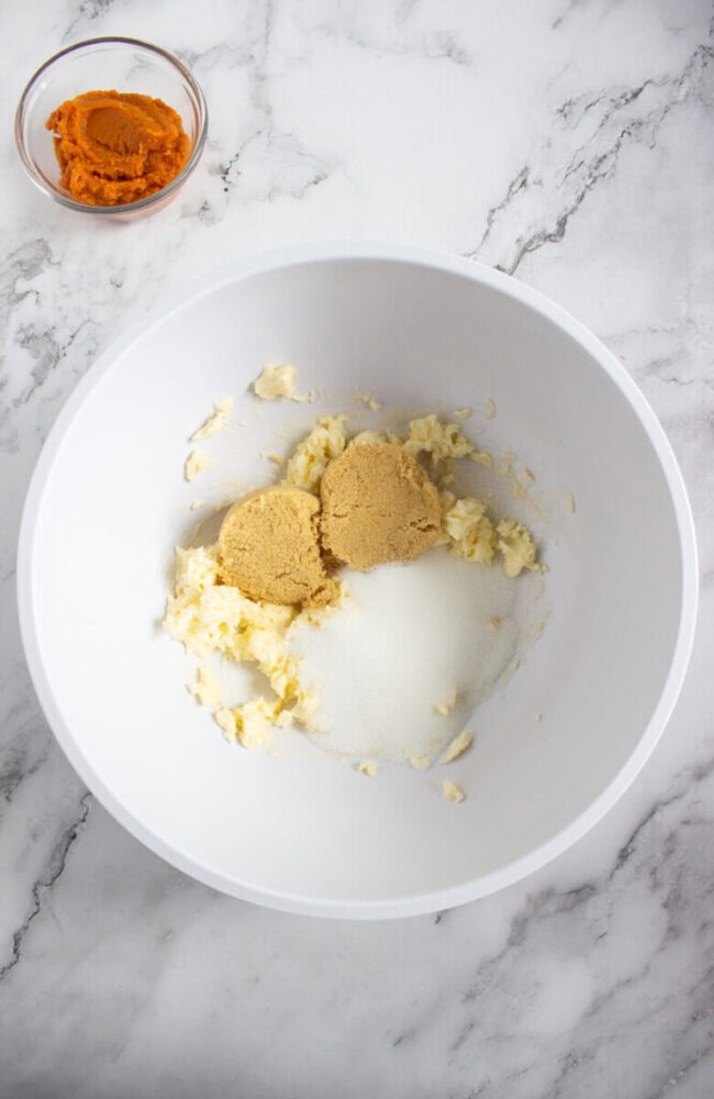 A white mixing bowl with softened butter, brown sugar, and granulated sugar, next to a small bowl of pumpkin puree on a marble countertop.
