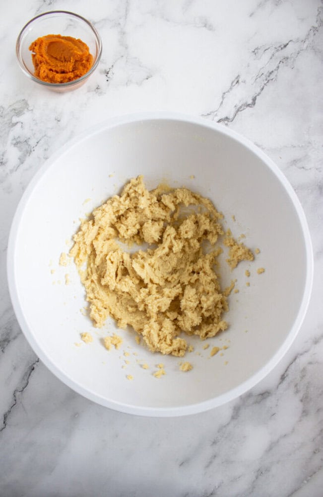 A white bowl with cookie dough mixture on a marble surface, with a small glass bowl of pumpkin puree in the background.