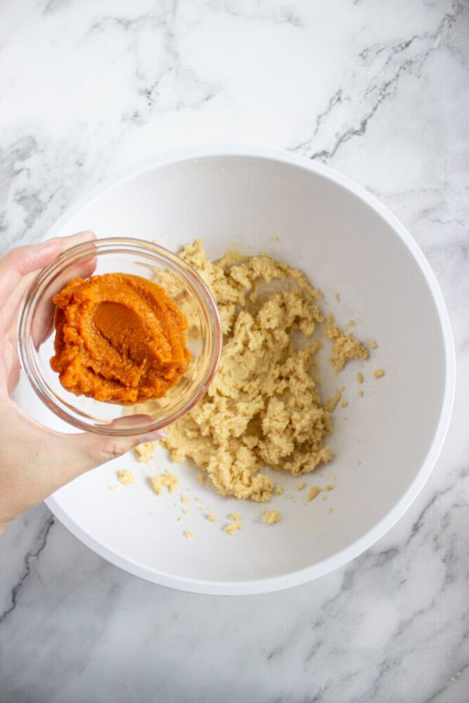 A hand holds a small bowl of pumpkin puree above a white mixing bowl containing a creamed mixture on a marble countertop.
