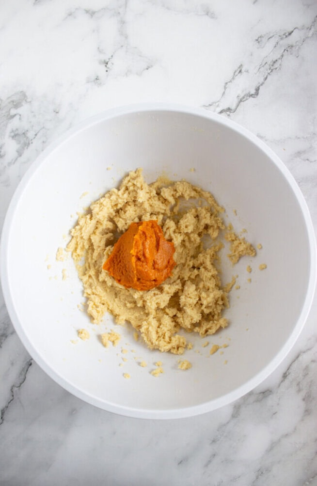 A white mixing bowl on a marble surface contains a partially mixed dough and a dollop of pumpkin puree on top.